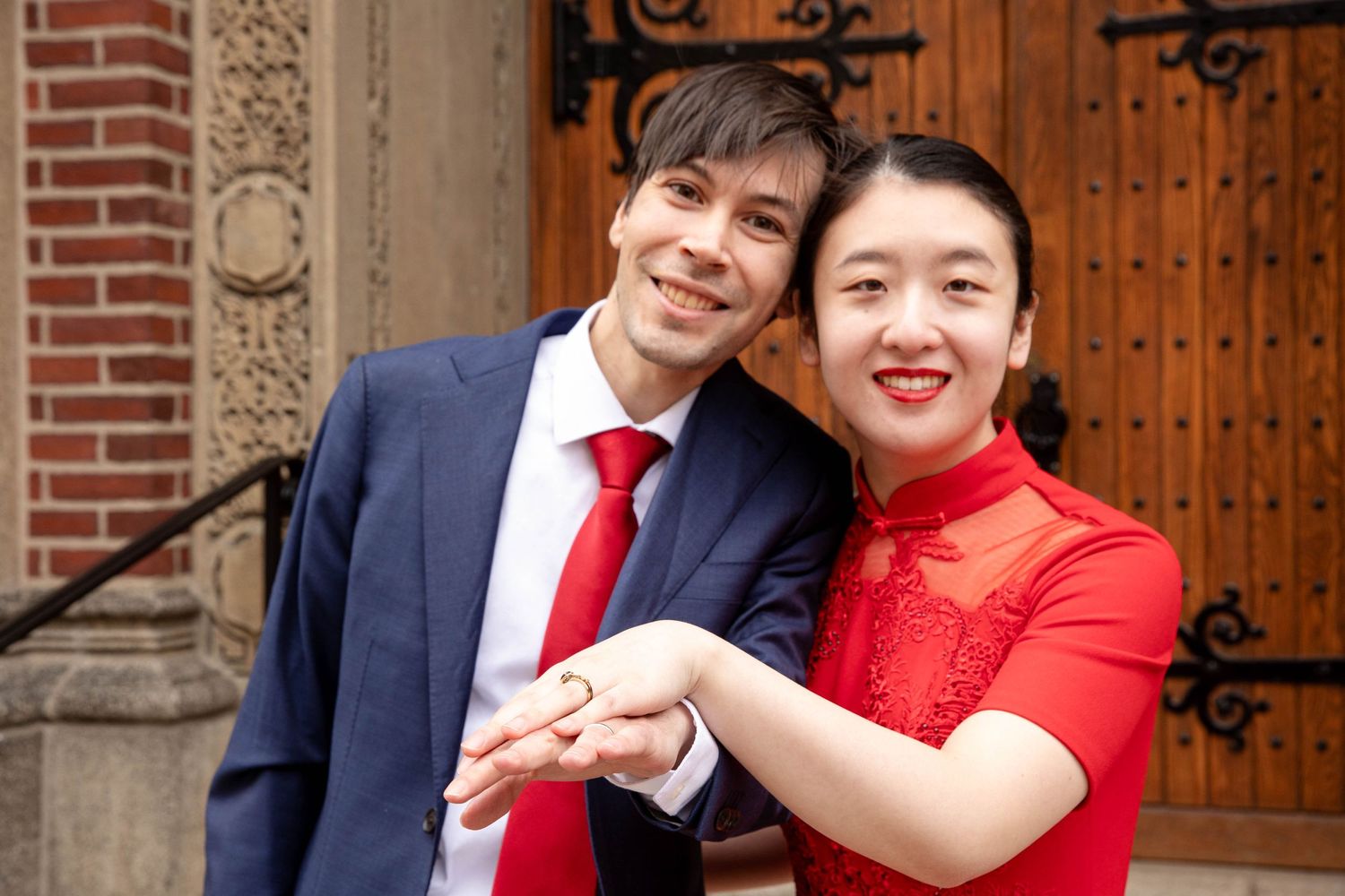 A couple poses for wedding photos outside a church door wearing red and navy formal attire.