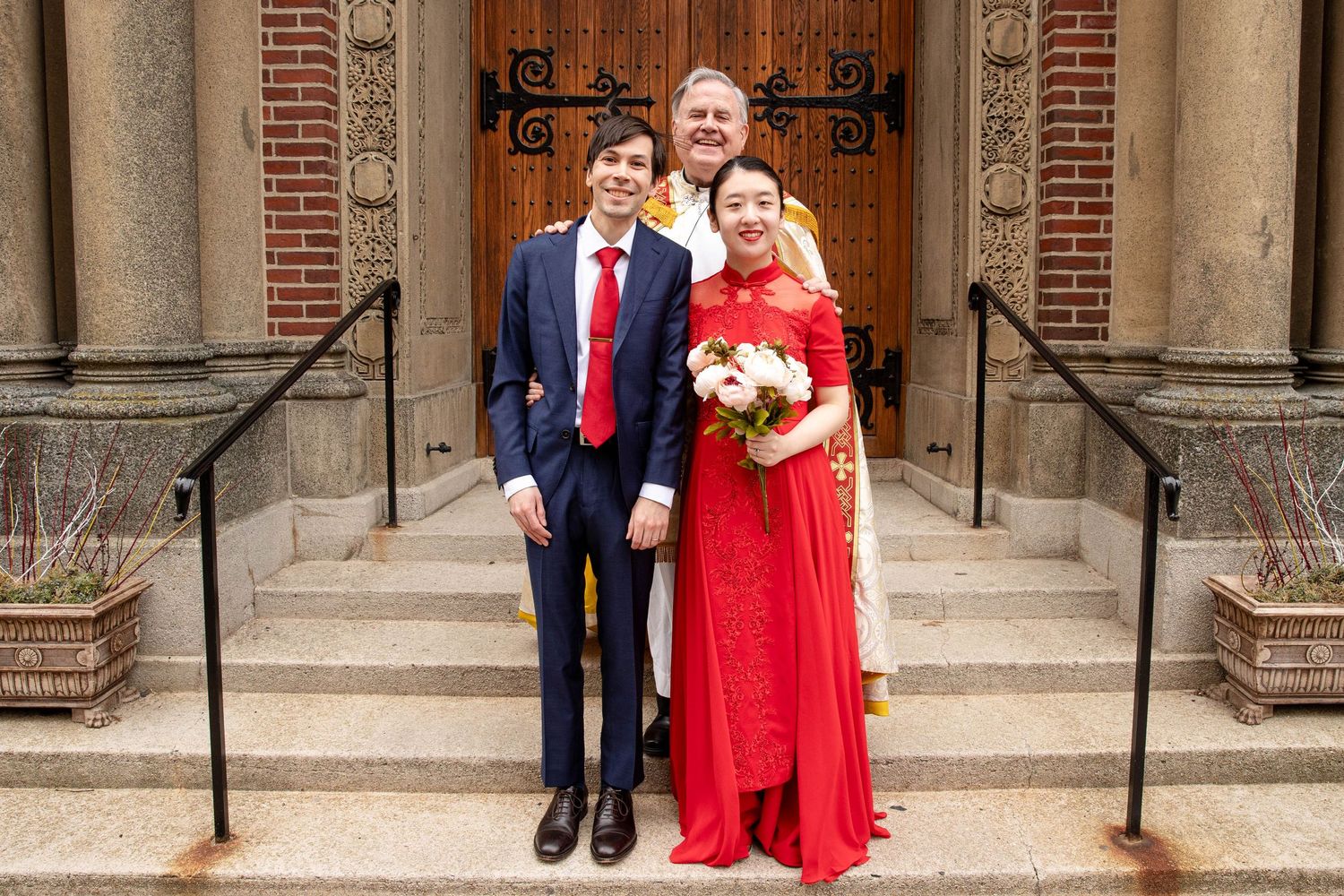 A wedding party of three poses on church steps with ornate wooden doors in the background.