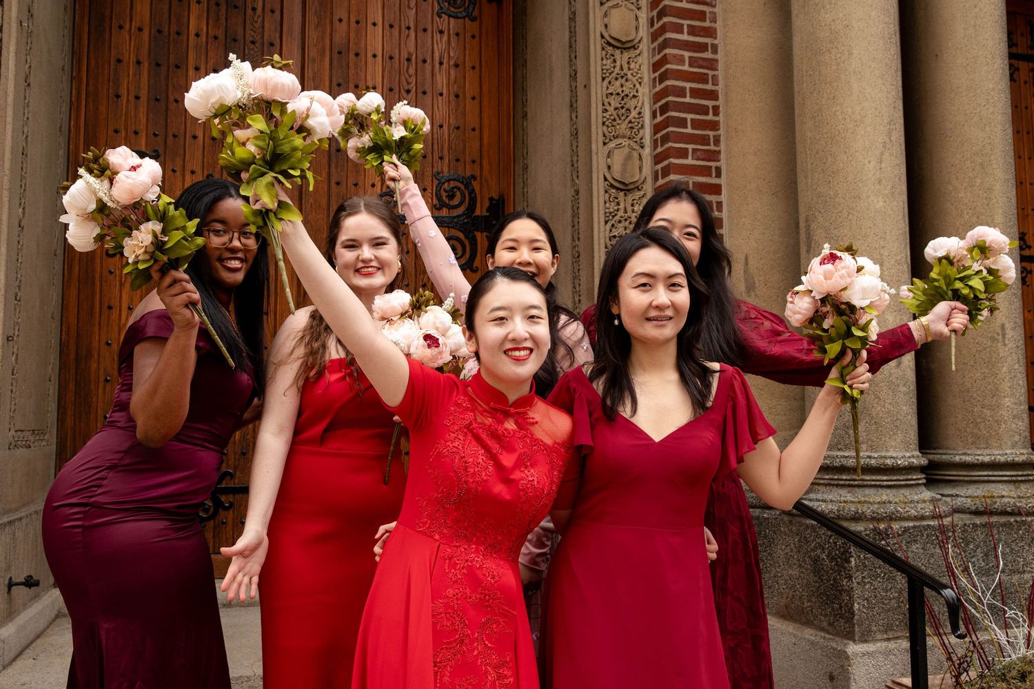 Bridesmaids in red and burgundy dresses hold bouquets while celebrating outside a church.