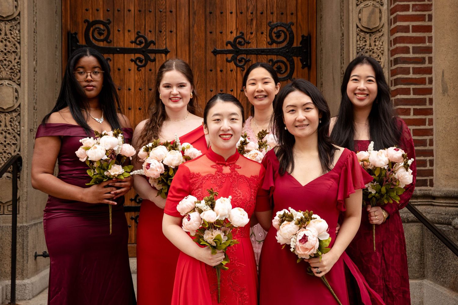 Bridesmaids in burgundy dresses pose with the bride in red holding floral bouquets.