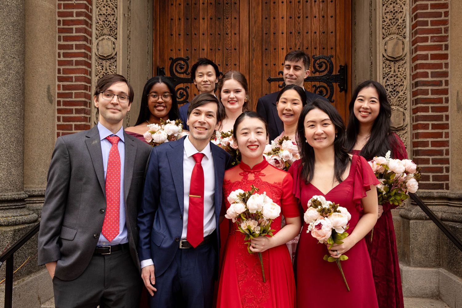 Wedding party members in red and burgundy attire gather for formal photo in front of church entrance.