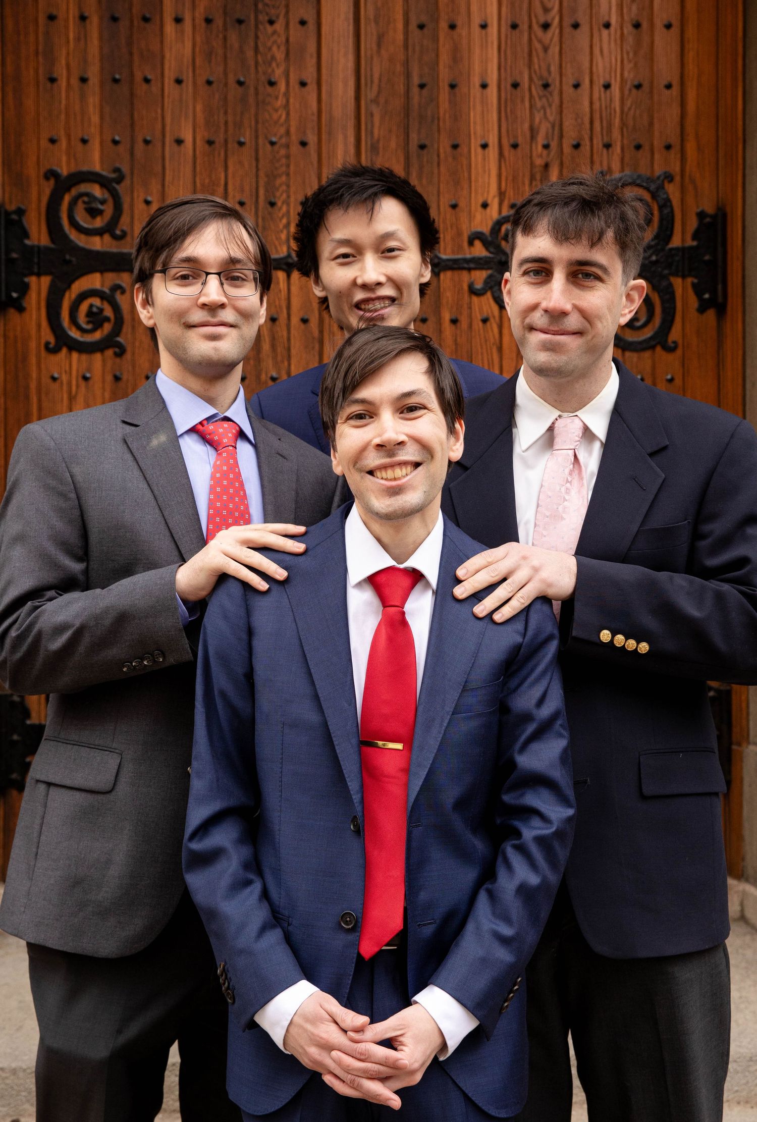 Groomsmen in suits and red ties pose playfully in front of wooden church doors.