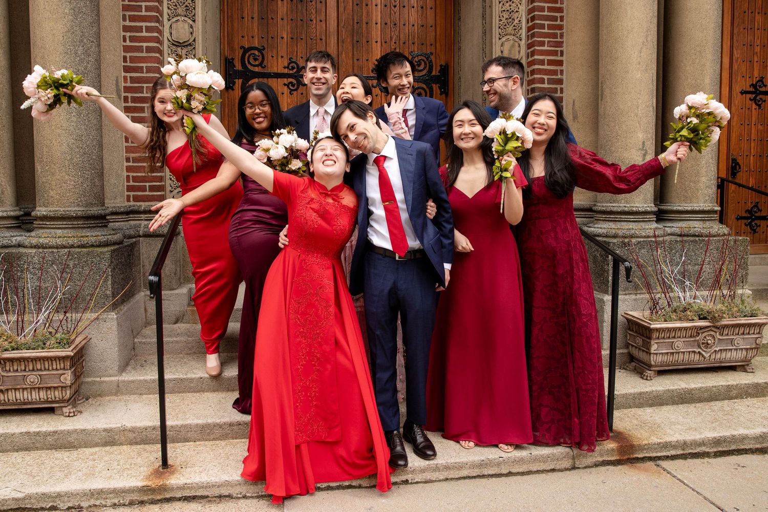 Wedding party strikes fun pose with arms outstretched holding bouquets in front of church doors.