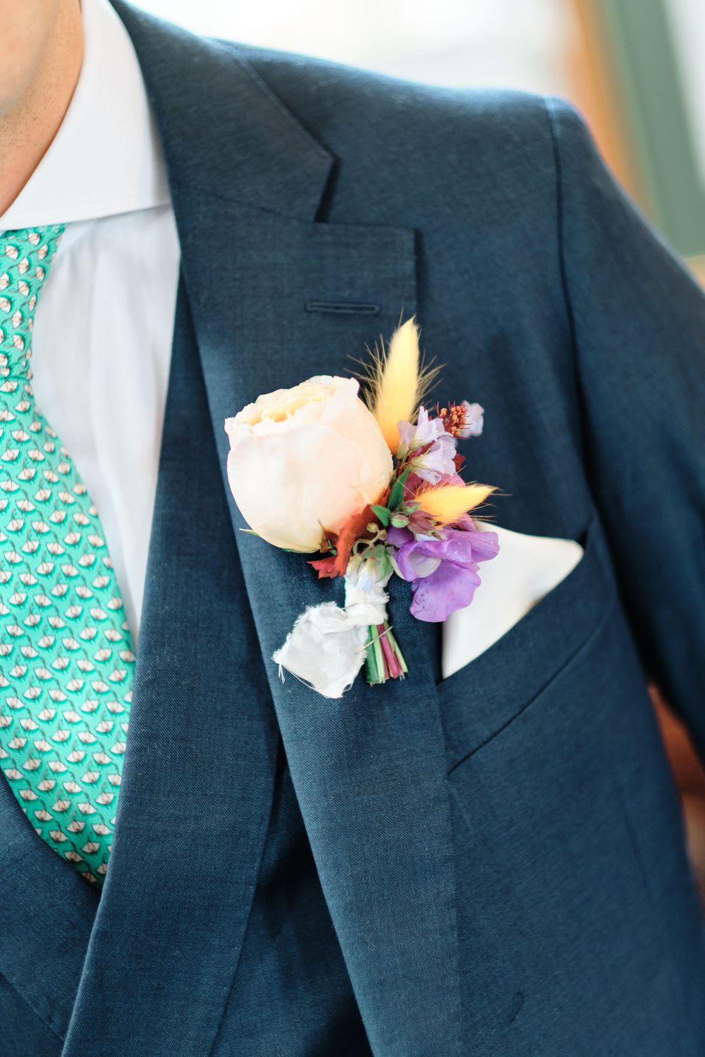 A delicate boutonniere with peach rose and purple flowers on a navy suit lapel.