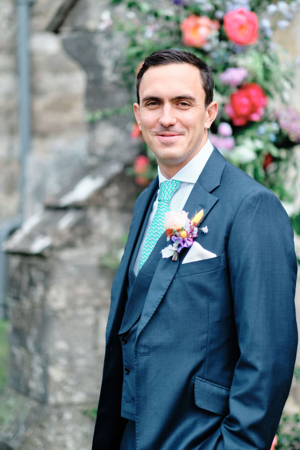 A groom in a navy suit with turquoise tie stands by floral decorations.