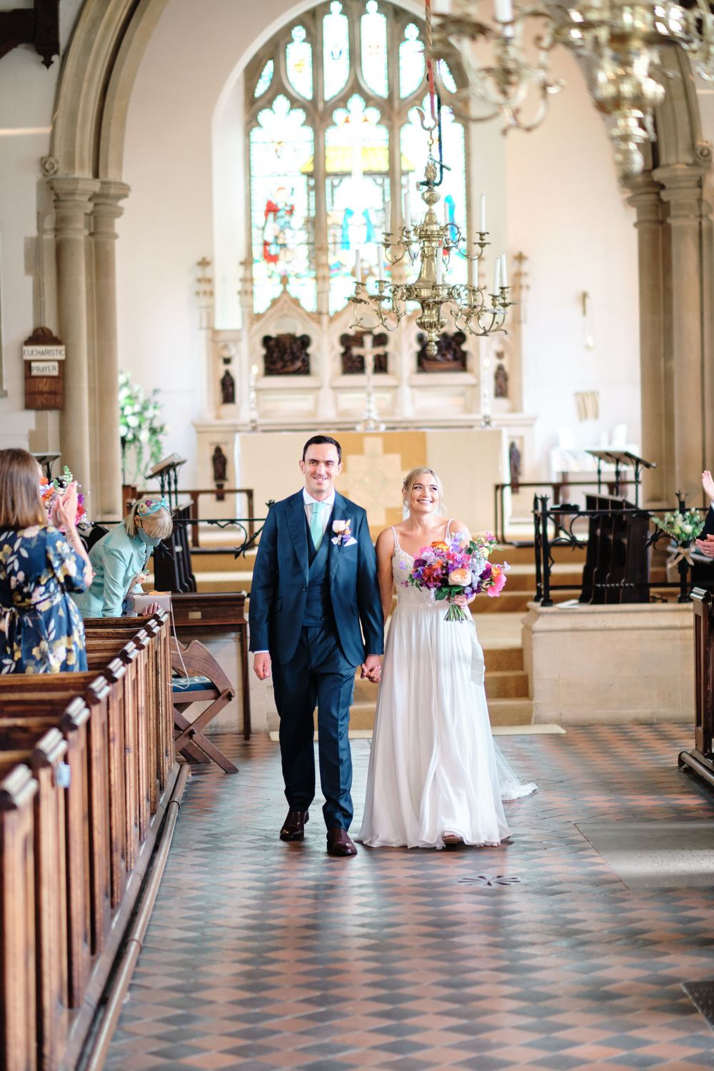 A couple walks down the church aisle with stained glass windows and chandelier overhead.