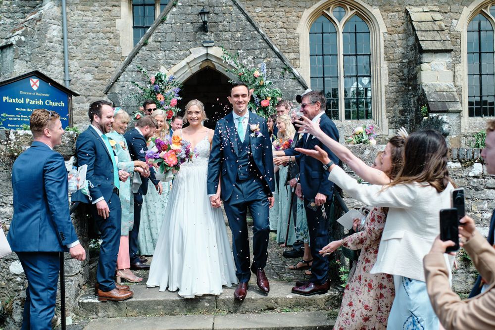 Wedding guests throw confetti as couple exits stone church building.