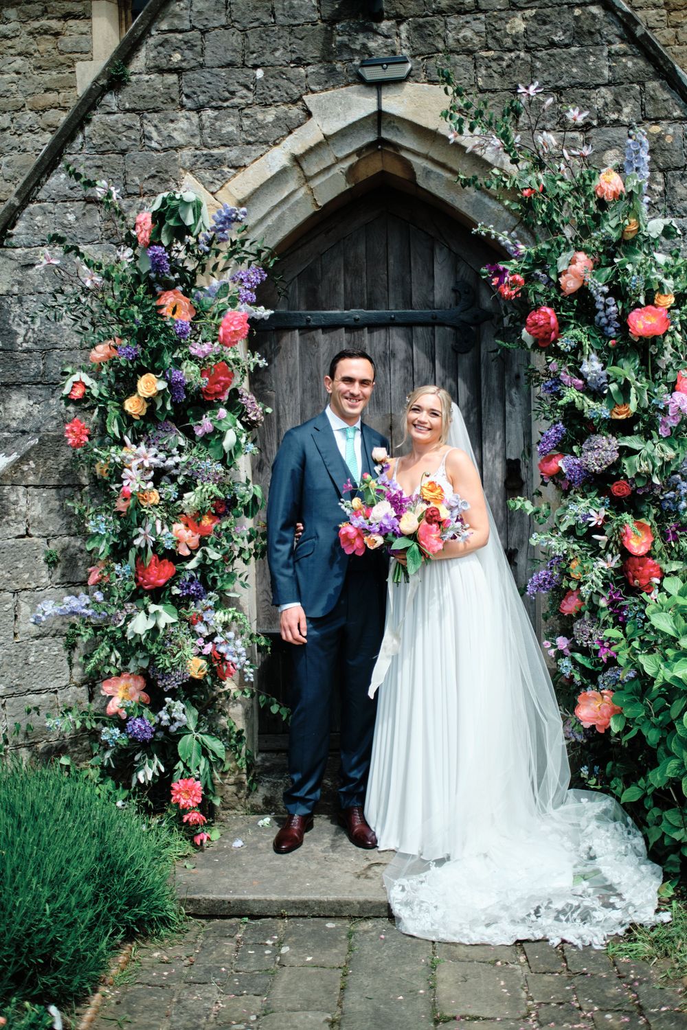 A romantic wedding photo in front of a stone church doorway adorned with colorful floral arch arrangements.