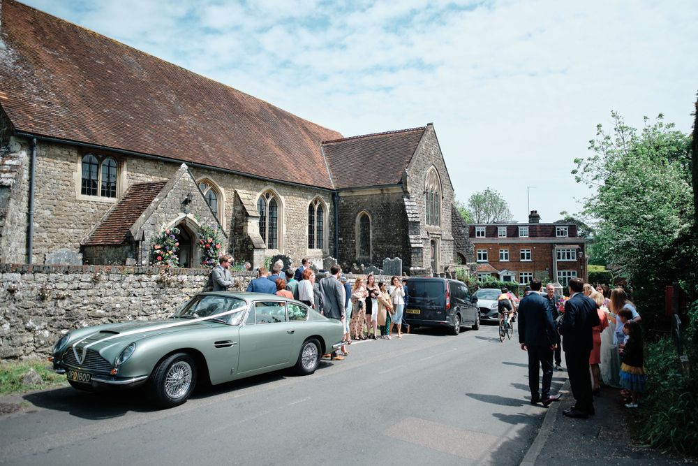 Wedding guests gather outside a historic stone church as a classic silver Aston Martin awaits.