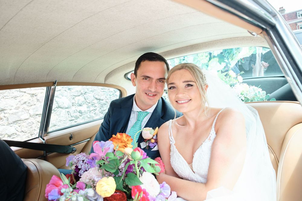 Wedding couple sitting in the back of a vintage car with colorful floral bouquets.