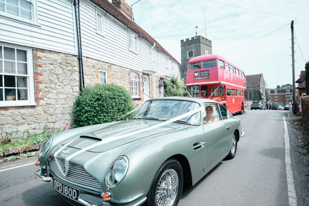 Classic silver Aston Martin DB5 parked on a village street with a red double-decker bus in background.