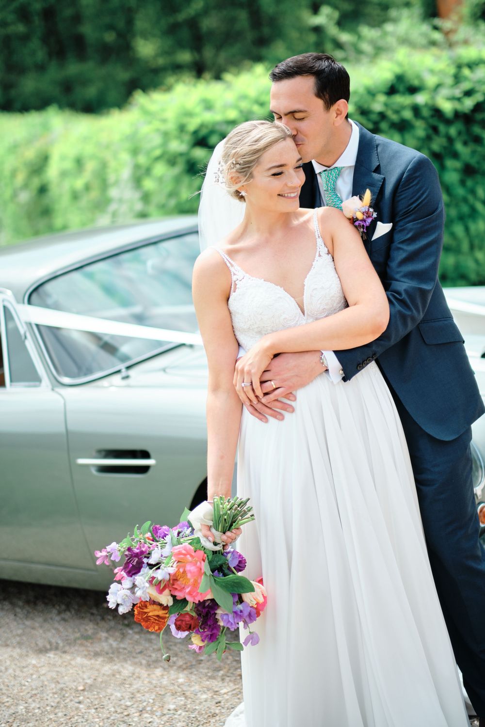 A romantic wedding couple embrace next to a vintage car with a colorful bouquet of flowers.