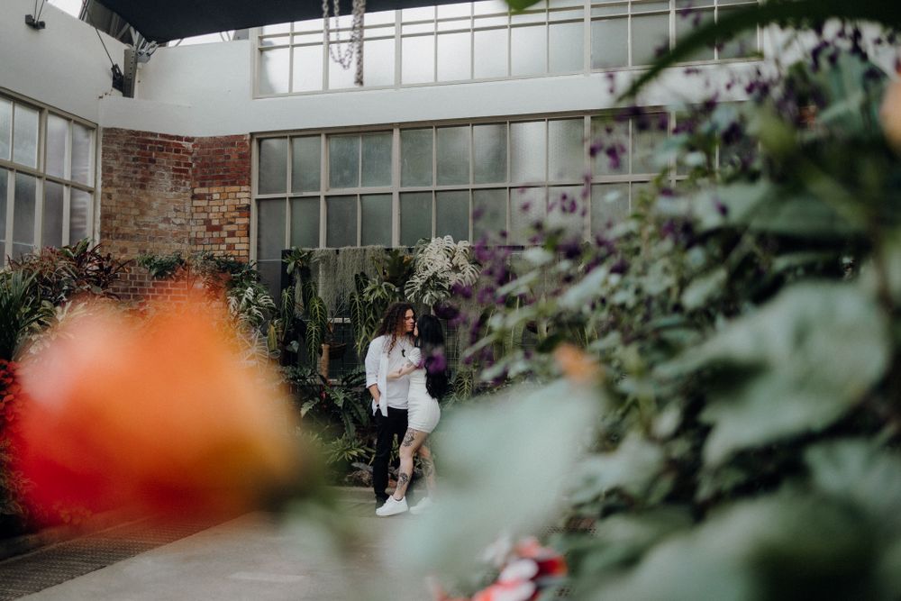 A couple embraces in a greenhouse setting with brick walls and flowering plants.