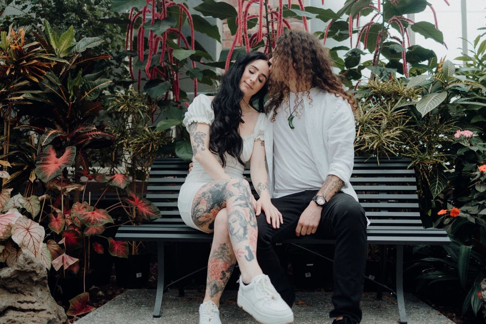 Two people sit together on a black bench surrounded by lush greenery and red flowering plants.
