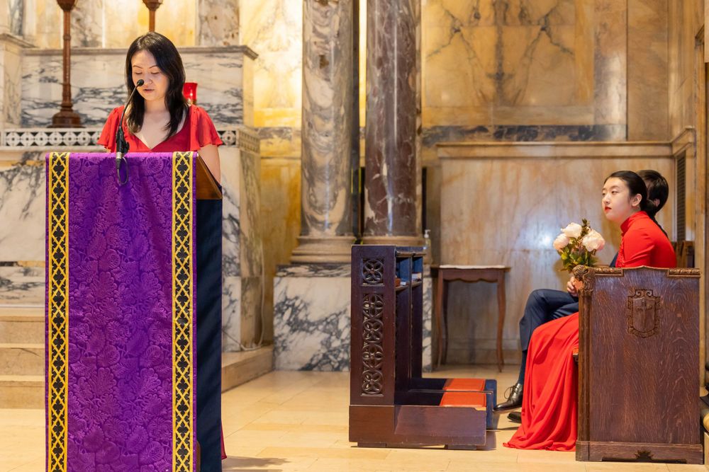 People speaking at a church lectern with purple cloth and ornate stone walls in the background.