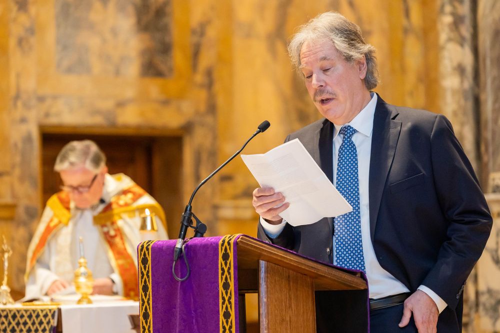 Someone in a navy suit reads from a podium with a purple cloth in an ornate church setting.