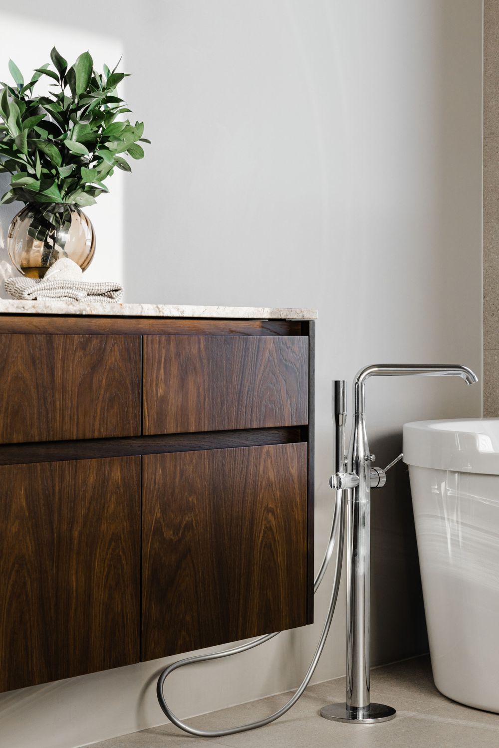 Elegant freestanding bathtub faucet beside wooden vanity with potted plant on marble top.