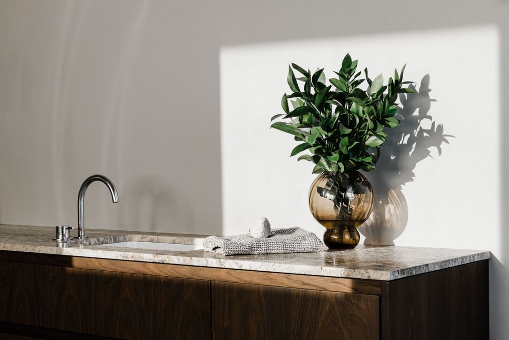 Sunlit wooden vanity with marble top and decorative vase containing green plants.