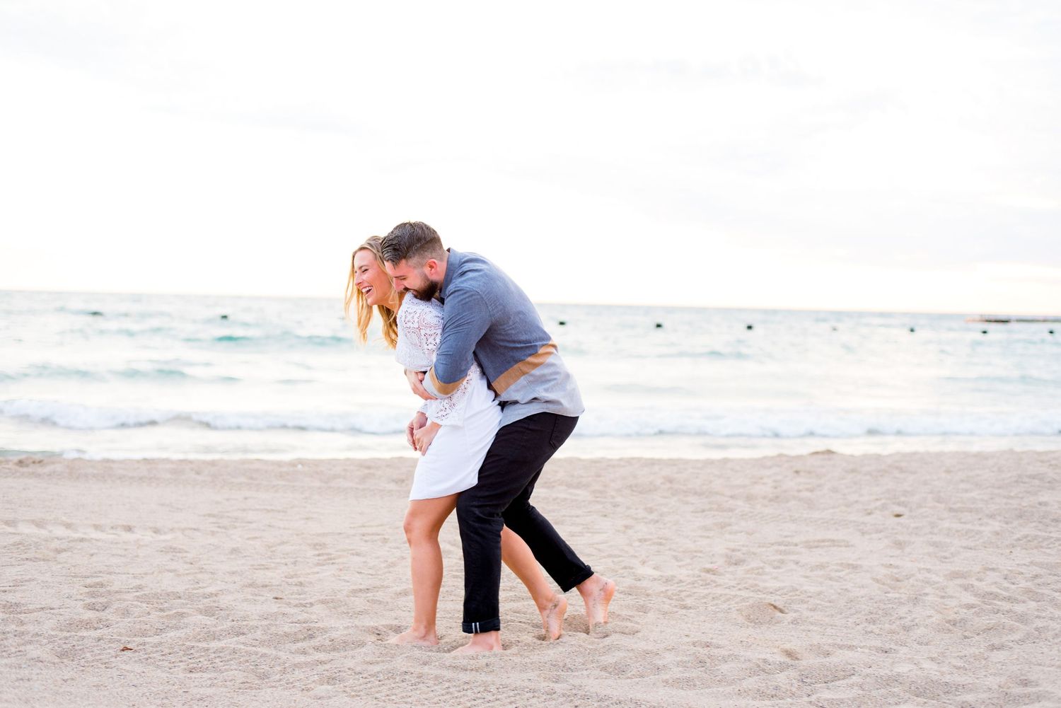 man with dark hair hugs woman with blonde hair in white lace dress from behind on North Avenue Beach