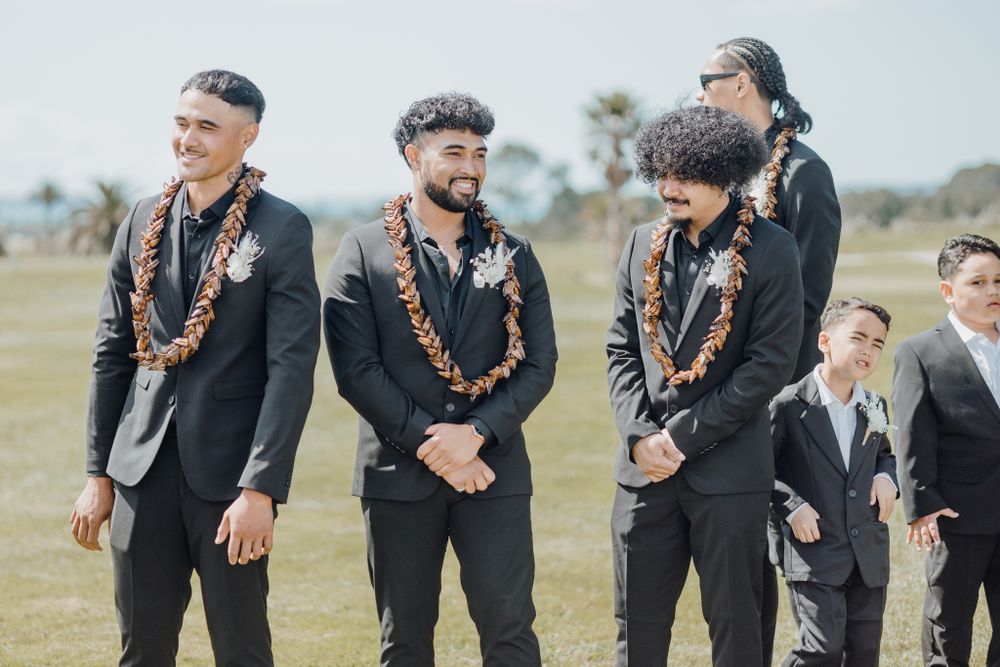 Groomsmen wearing black suits with wooden leis smiling during an outdoor wedding ceremony.