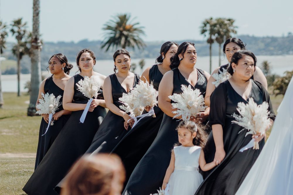 Bridesmaids in black dresses carrying white pampas grass bouquets at an outdoor wedding.