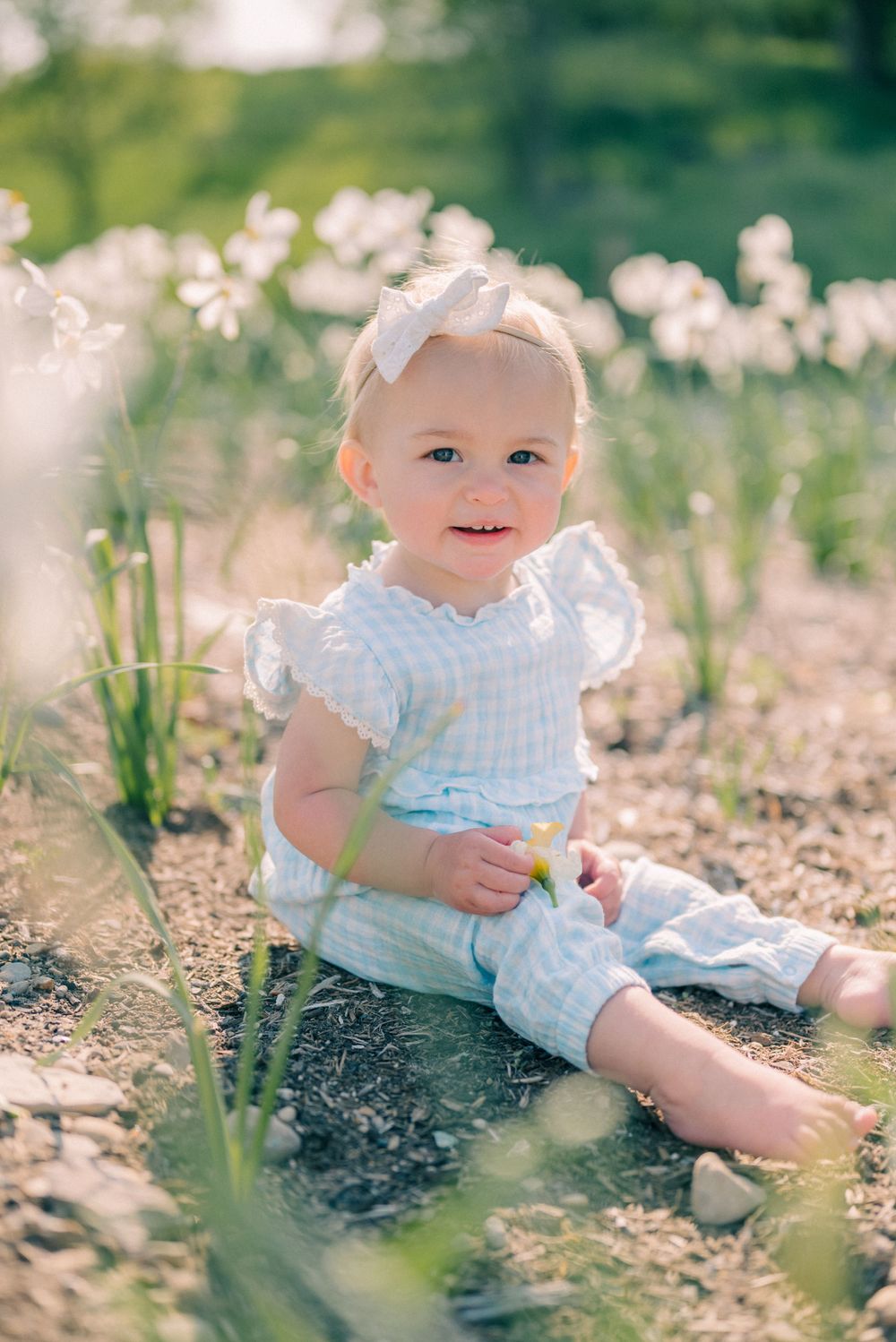 A small child in blue gingham outfit sits among spring flowers wearing a white bow.