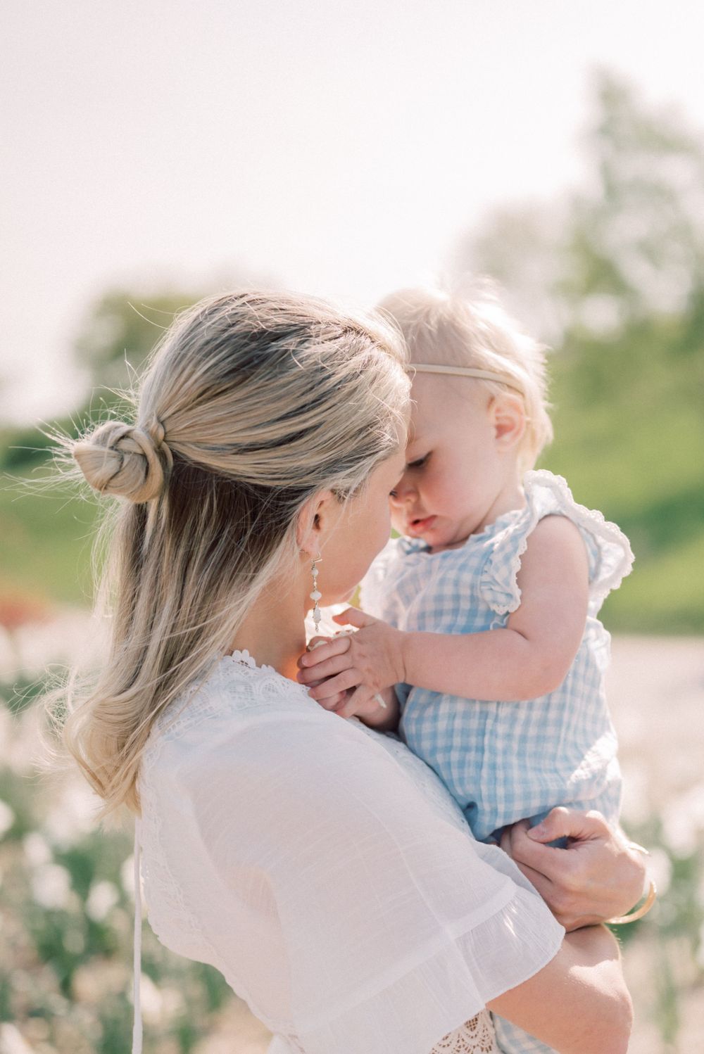 Tender outdoor moment between parent and baby in white dress and blue gingham outfit among spring flowers.