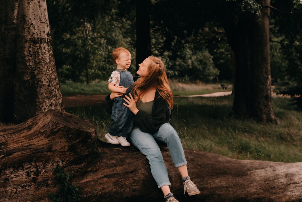 A parent and child share a joyful moment while sitting together on a tree trunk in a wooded park setting.