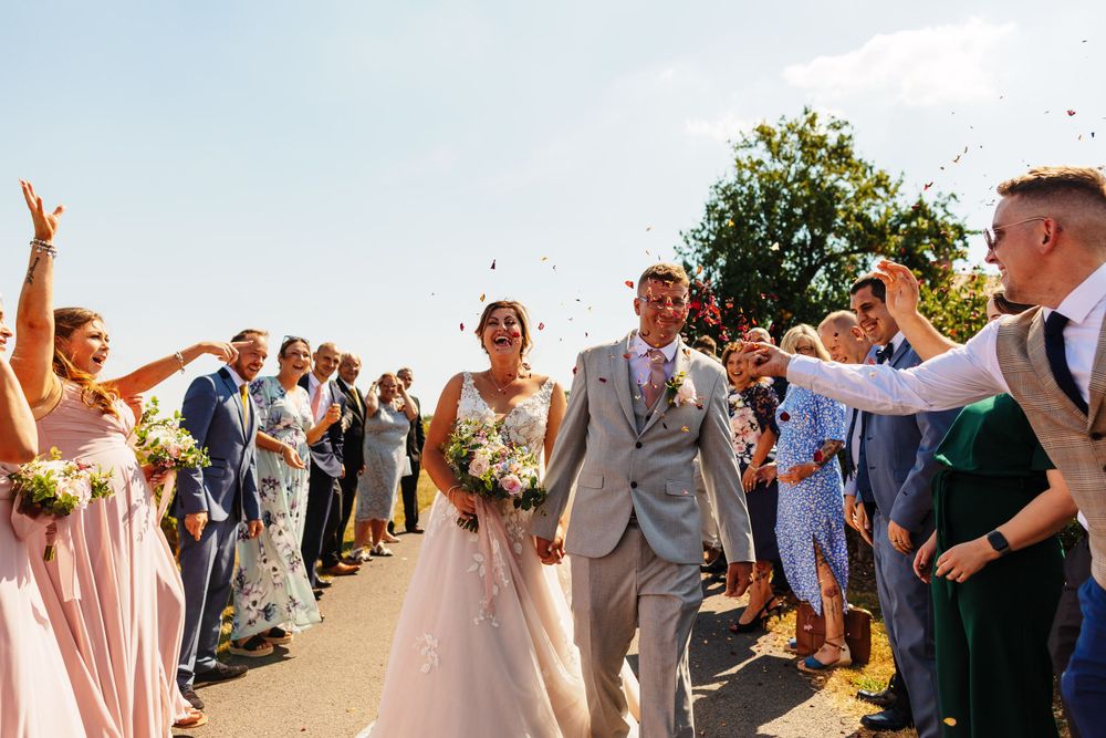 Wedding guests toss confetti over celebrating newlyweds on a sunny countryside path.
