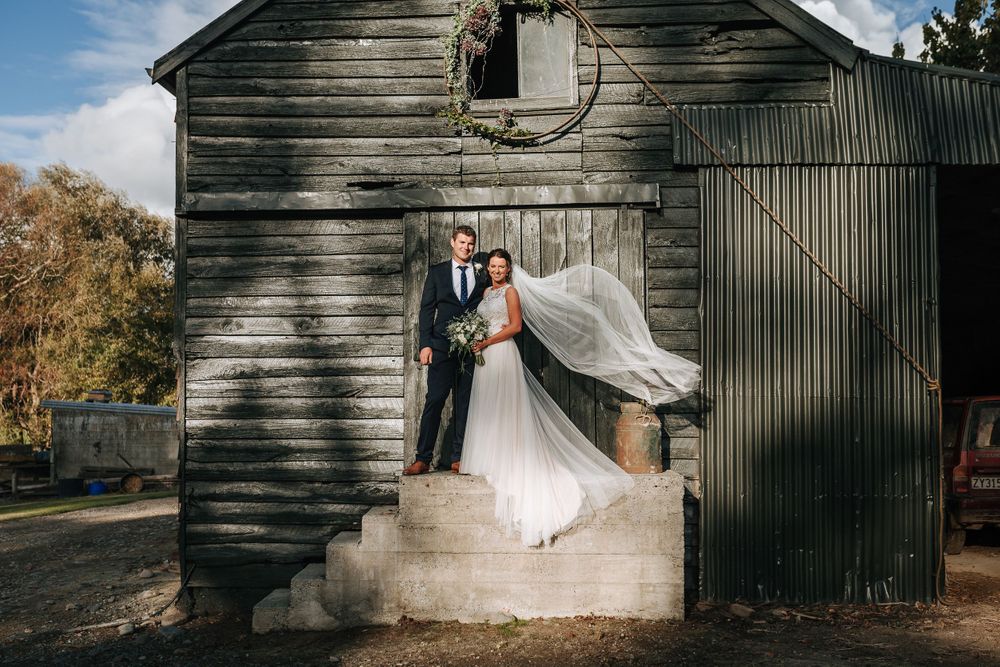 Bride and groom in front of black farm shed in Canterbury with veil blowing in air