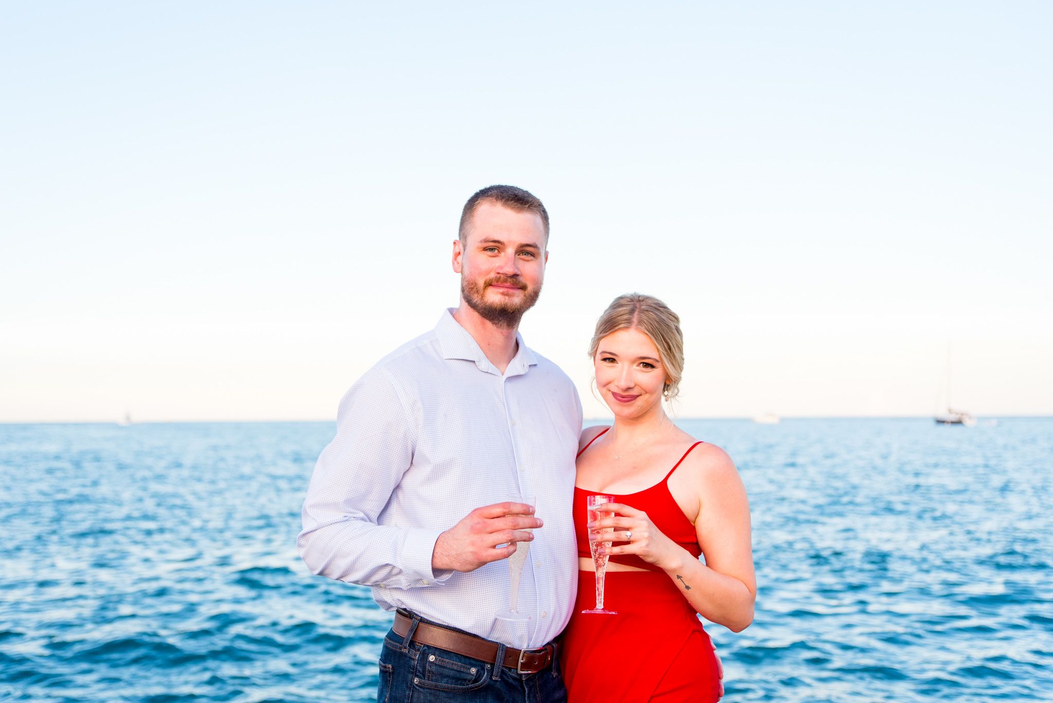 blonde woman in red dress and man in white shirt holding glasses of champagne in front of Lake Michigan in Chicago