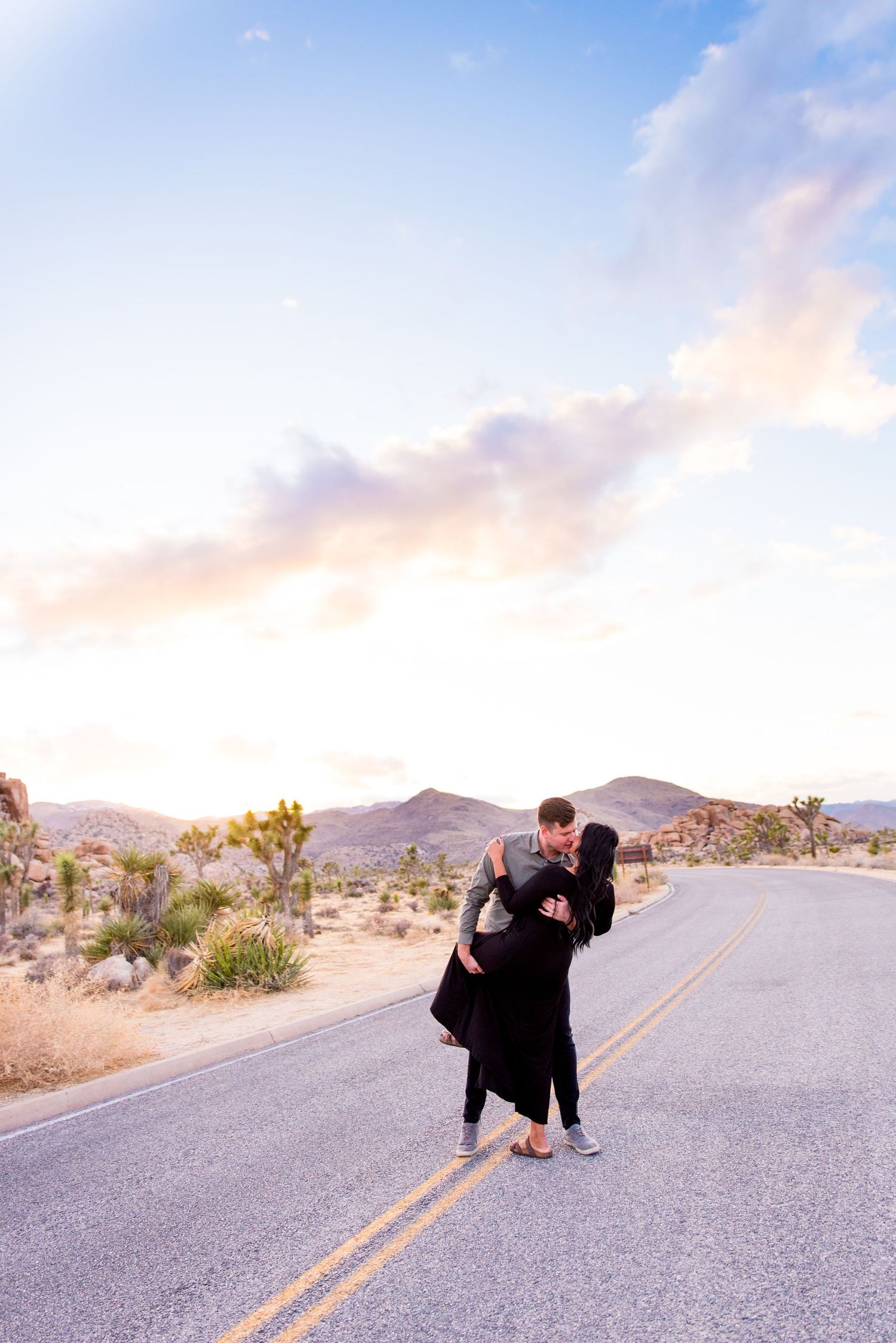 man in gray shirt dips his fiancee in black dress in the middle of the road during sunset at Joshua Tree National Park