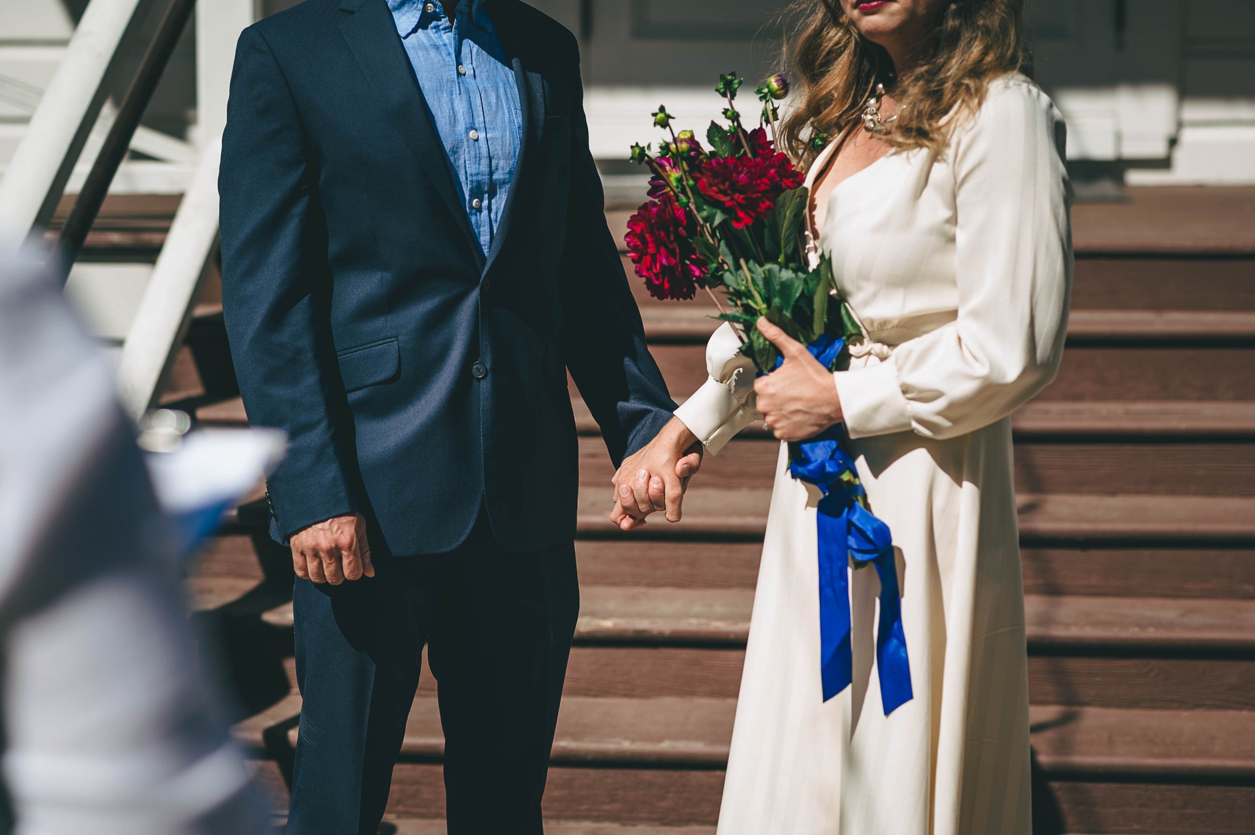 Detail of bride and groom holding hands and the brides bright red bouquet tied with a blue ribbon.