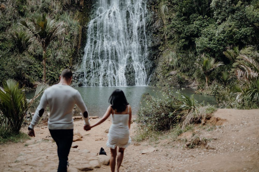 A couple holding hands while walking towards a majestic waterfall in a lush forest setting.