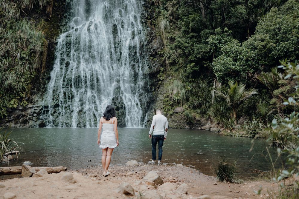 Two people standing at the base of a tall cascading waterfall surrounded by tropical vegetation.