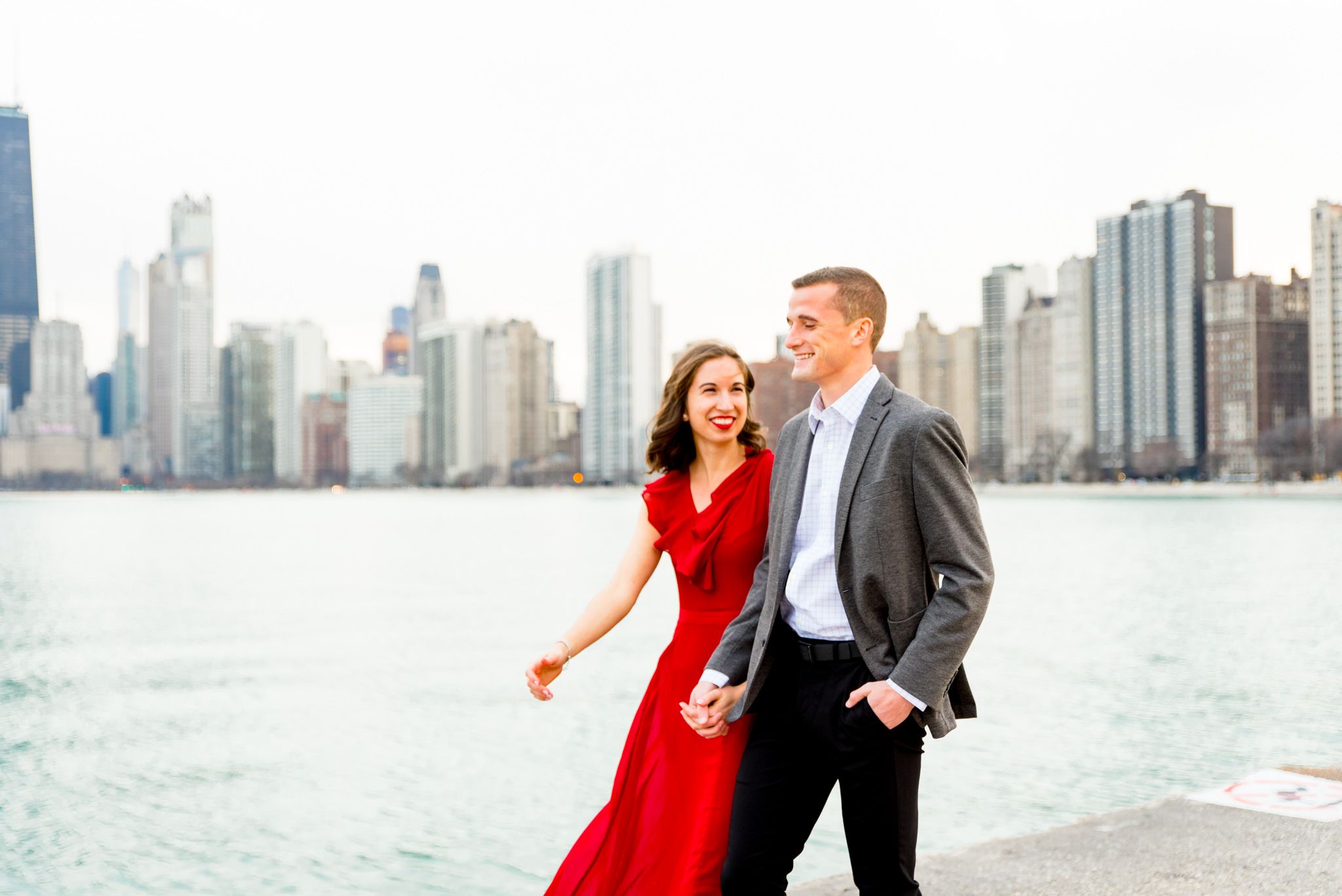 woman in red dress holding hands walking with man in grey suit along North Avenue Beach Chicago engagement pictures