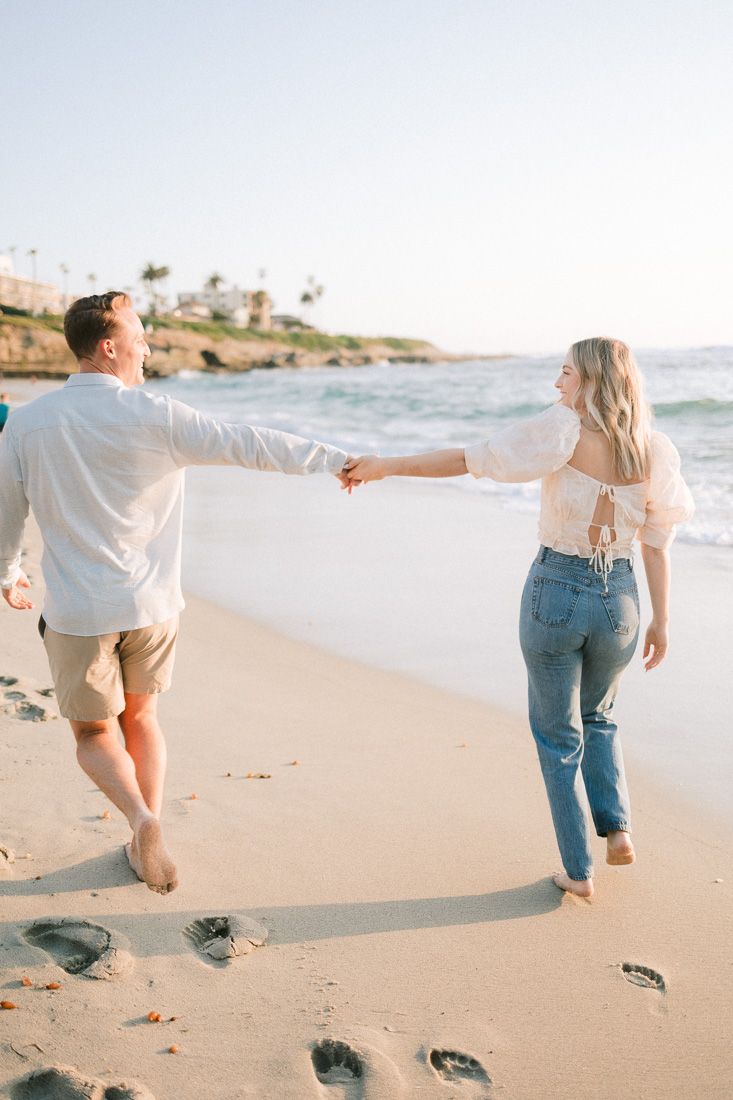 Couple holding hands and walking on a sunny beach with coastal cliffs in the background.