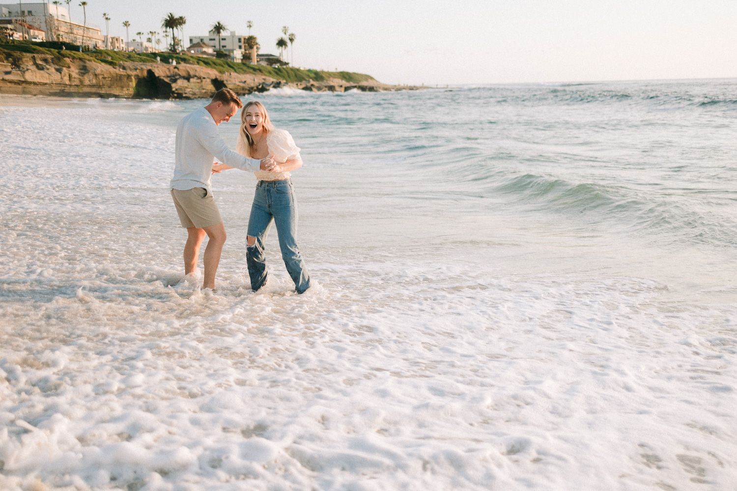Couple playing in the ocean waves at sunset with coastal cliffs visible.
