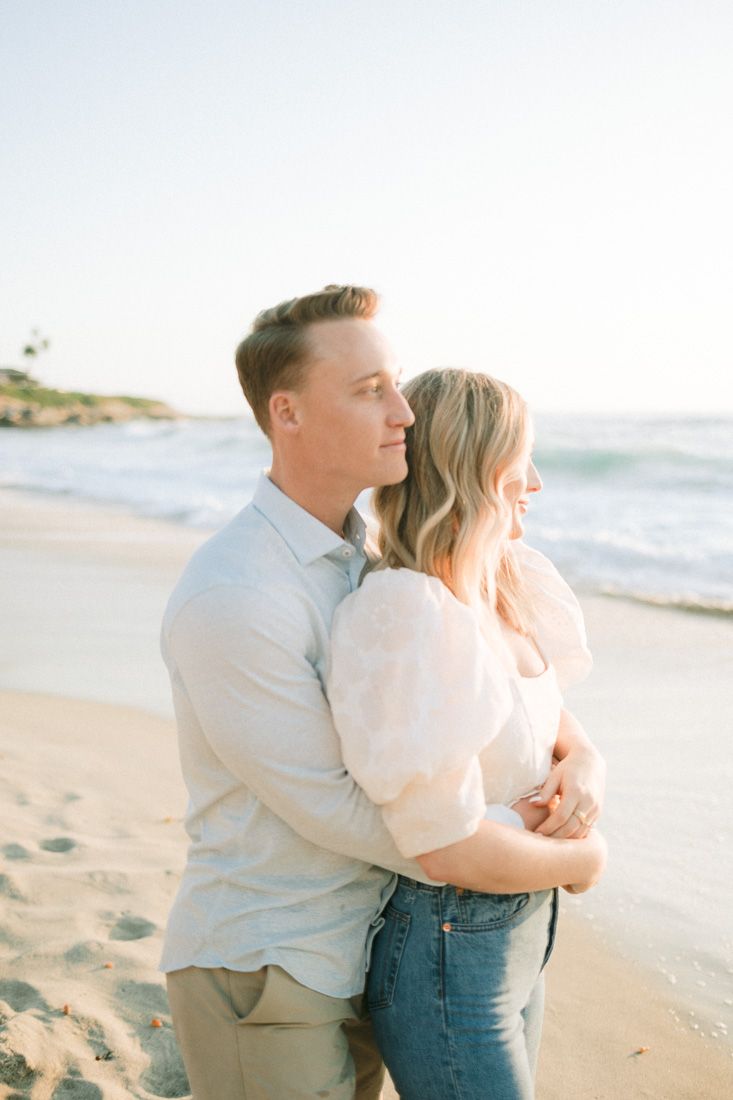 Couple embracing on a sunny beach with lighthouse and ocean waves in the background.