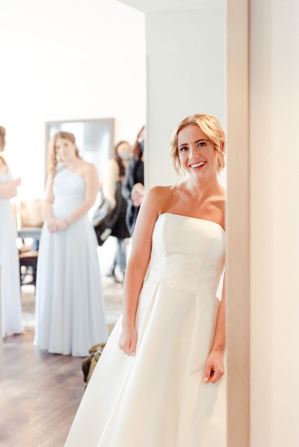 blonde bride with updo smiling at herself in the mirror as bridesmaids watch from the background