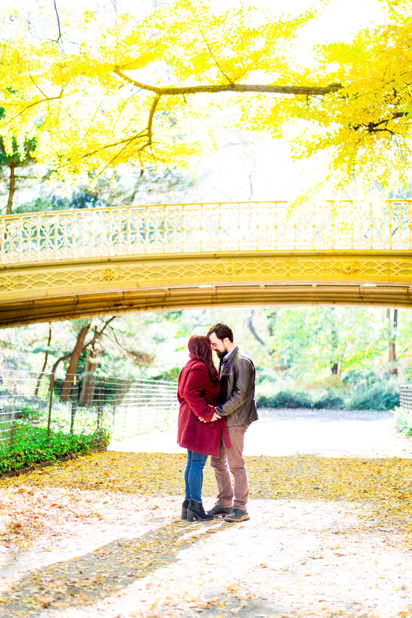 man in brown leather jacket holding hands with woman in red jacket under Pinebank Arch at Central Park in the fall