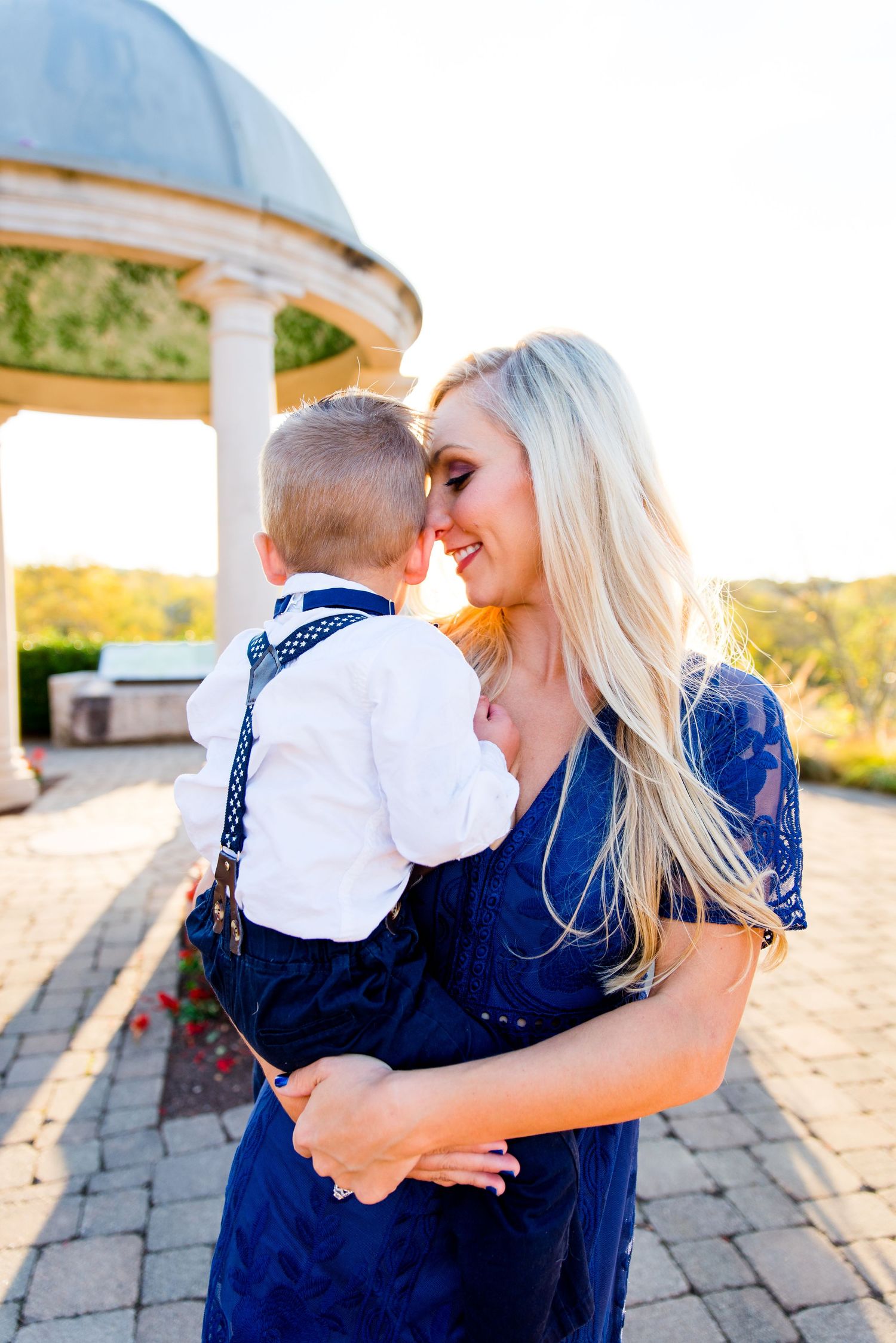 mom with long blonde hair in a navy lace dress putting her nose on her young baby son's cheek