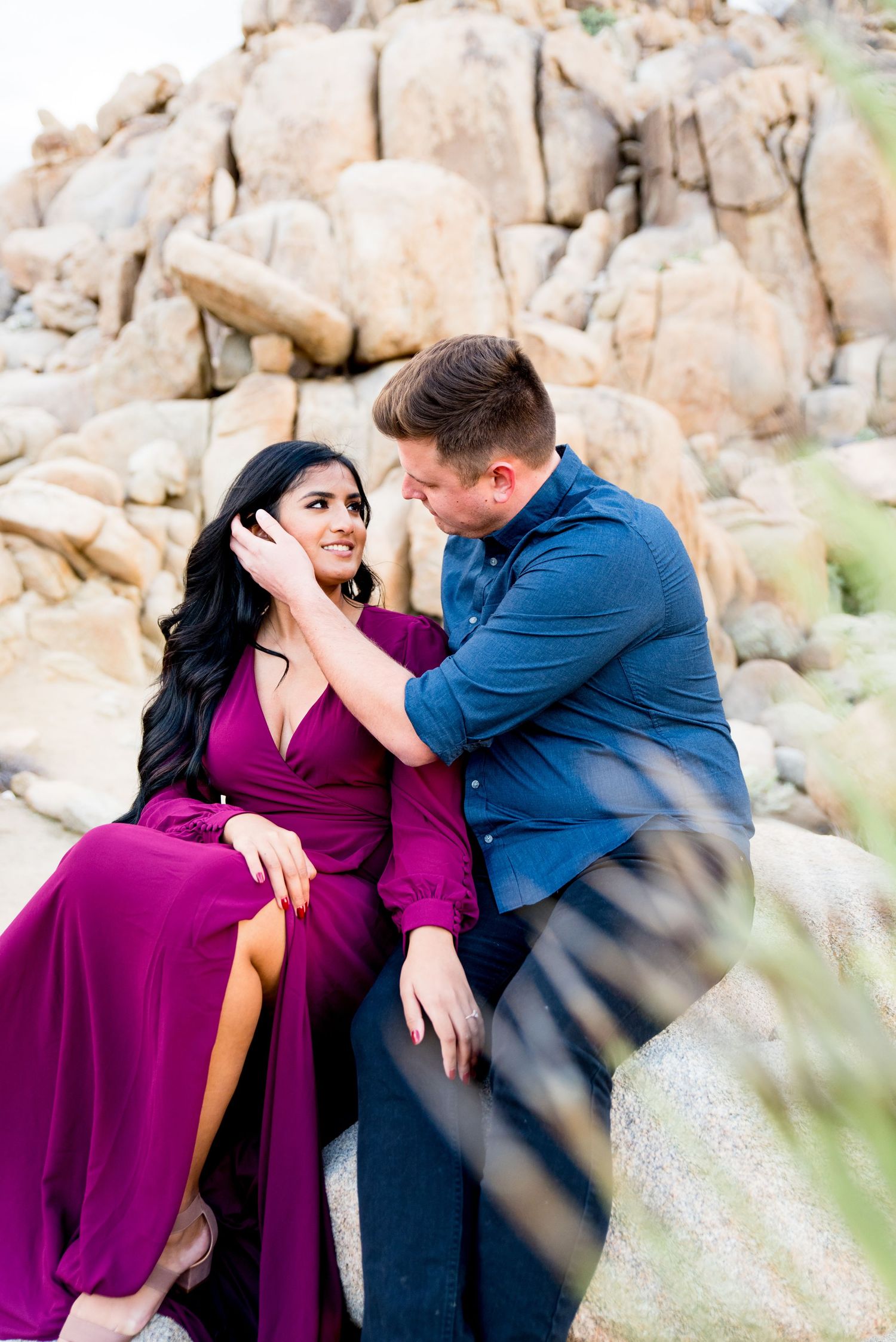man in blue button down puts hand on face of woman with long dark hair in a red dress at Joshua Tree Park in California