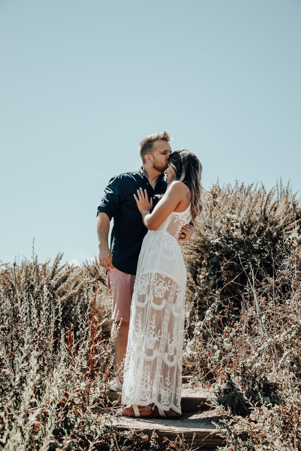 Romantic couple stands together among dried grasses during outdoor golden hour photoshoot wearing blue shirt and lace dress.