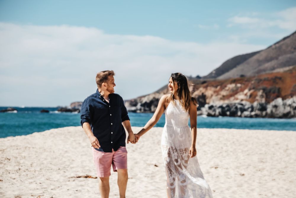 Couple holding hands while walking on scenic coastal beach with mountains in background.