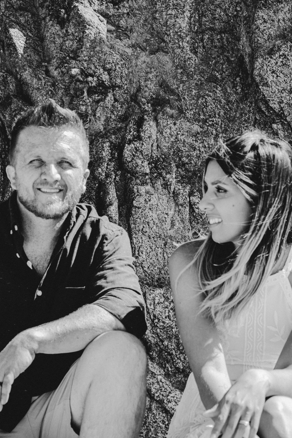 Black and white portrait of a couple smiling while sitting outdoors against a textured rock wall.