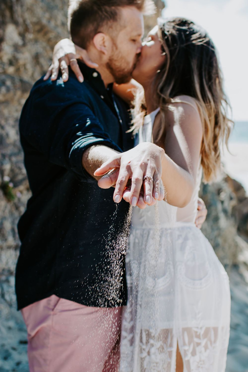 A romantic couple shares a kiss on a rocky beach while showing off an engagement ring in sparkling light.