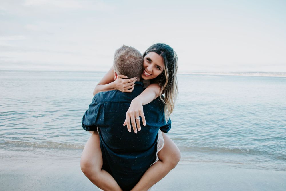 Two people embrace in a piggyback ride pose along a serene beach with calm ocean waters in the background.
