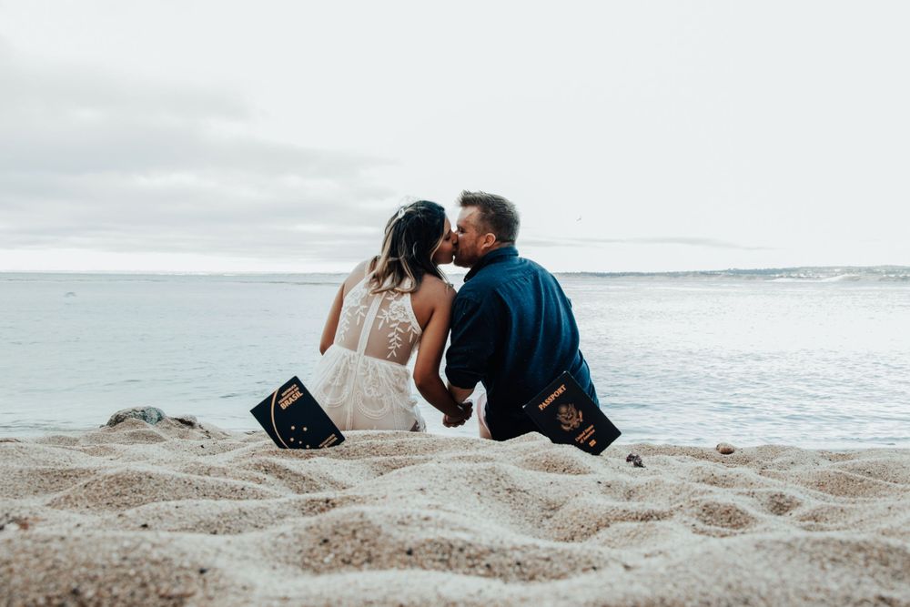 A couple in wedding attire sit close together on a sandy beach with the ocean and cloudy sky in the background.