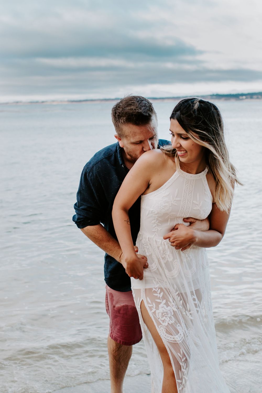 A romantic beach scene with a couple laughing together on the shoreline, she wearing a white lace dress against the ocean view.